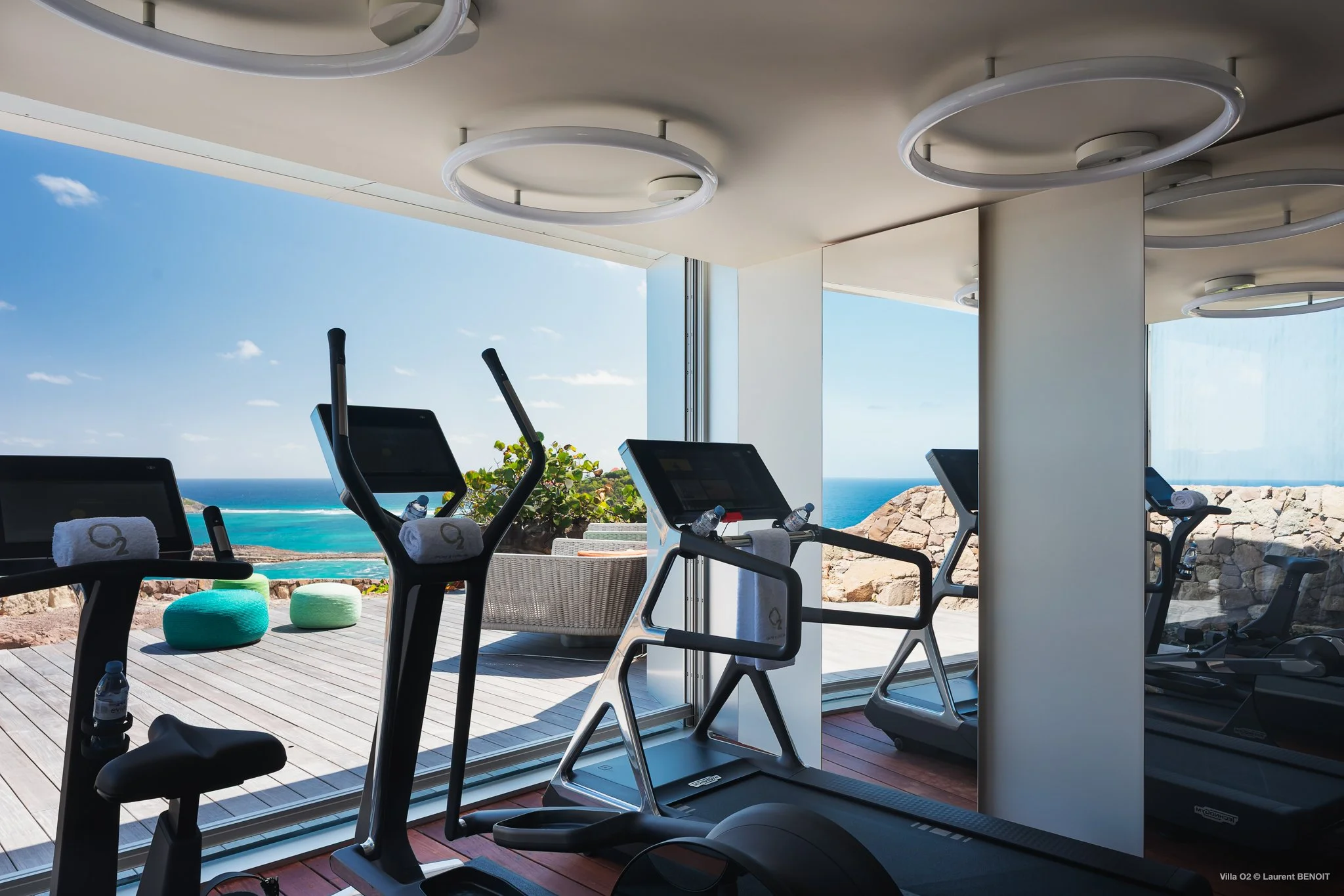 Modern outdoor patio with lounge seating, colorful ottomans, small round table, and a glass-enclosed gym area, with a stone wall and contemporary houses in the background.