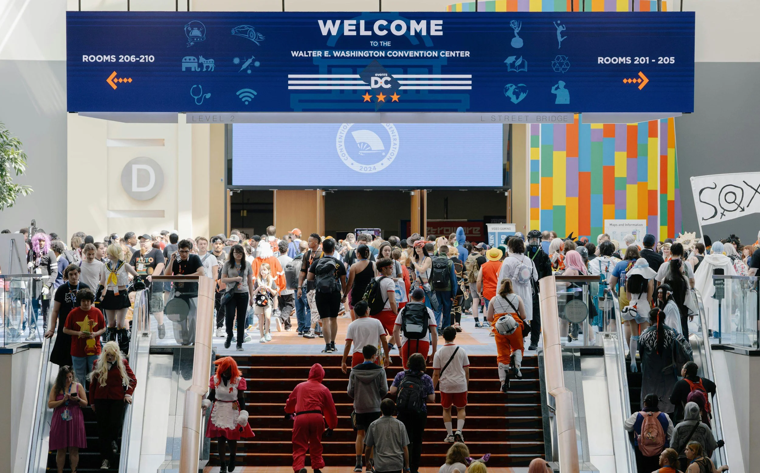 Crowd of people at a convention center with a large blue sign overhead that reads 'Welcome to the Walter E. Washington Convention Center.' The crowd includes individuals in casual clothing, some in costumes, and cosplayers. There are stairs and escalators leading to the entrance of the convention center.