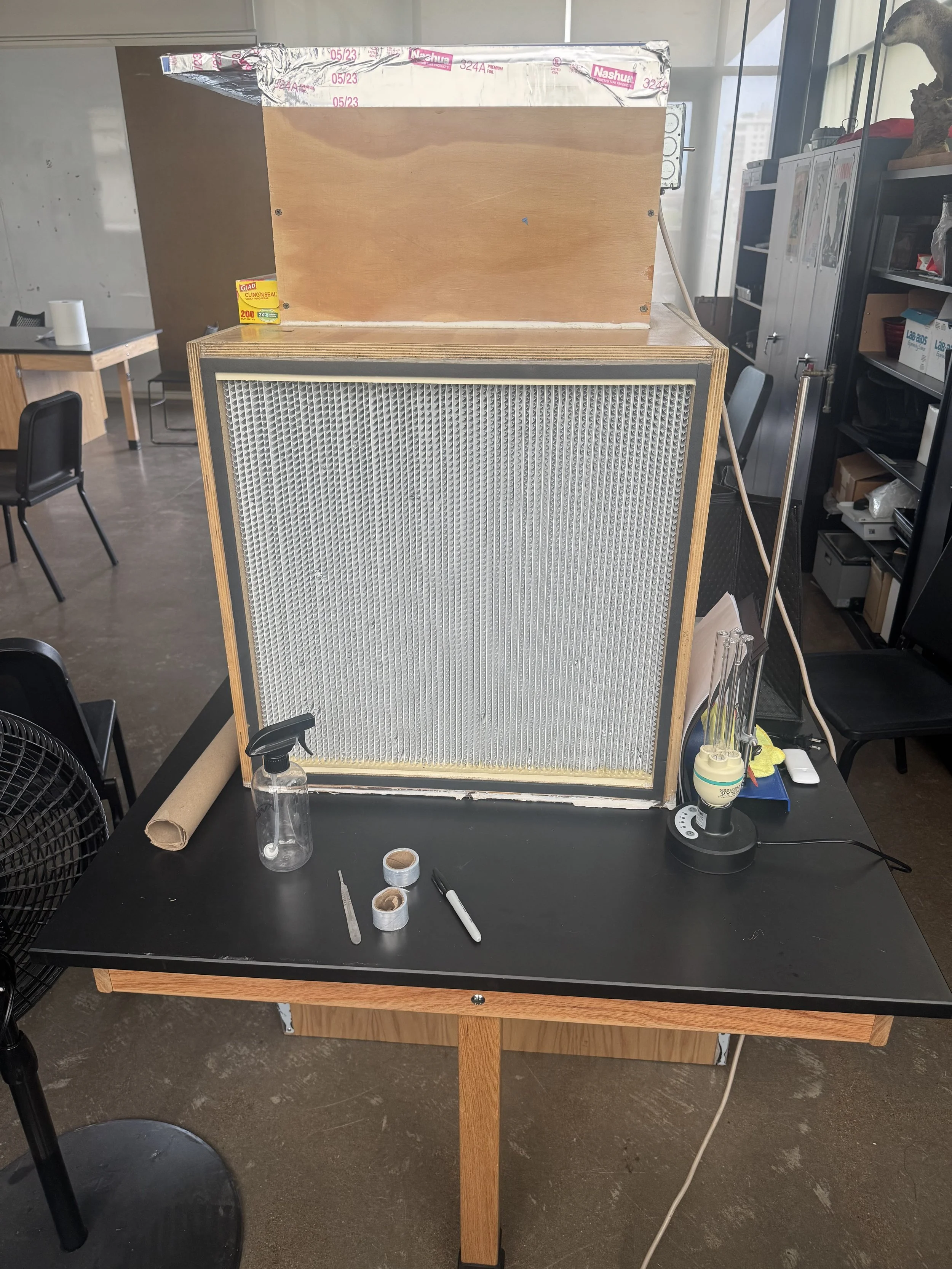 A workbench with various items including a small spray bottle, rolls of tape, a marker, and a test tube. Behind the workbench is a large device with a metal grille and a wooden top, resembling an experimental setup or a piece of lab equipment. The background shows a room with chairs, cabinets, and shelves with various objects.