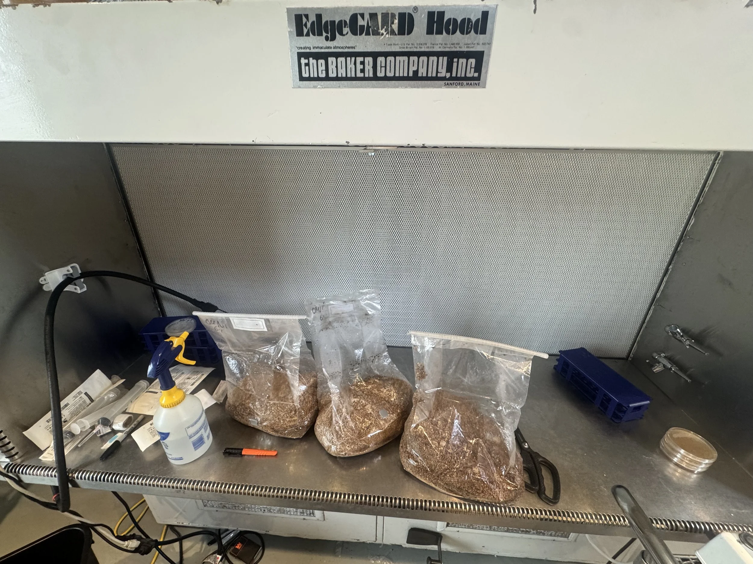 A metal surface with a sticker that reads 'EdgeCAMP Hood' and 'the BAKER COMPANY, inc.', along with a background of various tools, plastic bags, and supplies stored below the surface.
