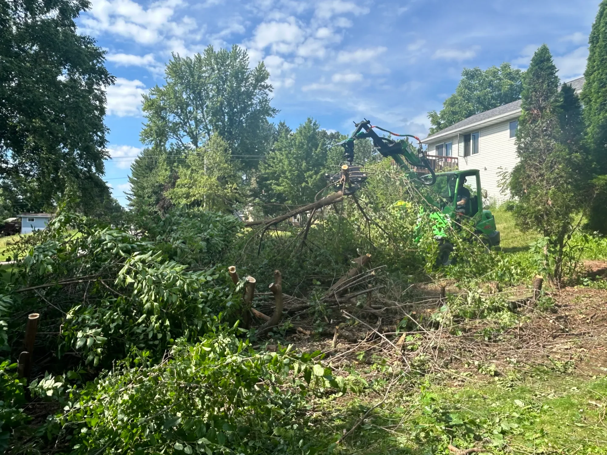 A small green tractor with a grapple attachment removing fallen tree branches in a backyard, with trees and a white house with a deck in the background.