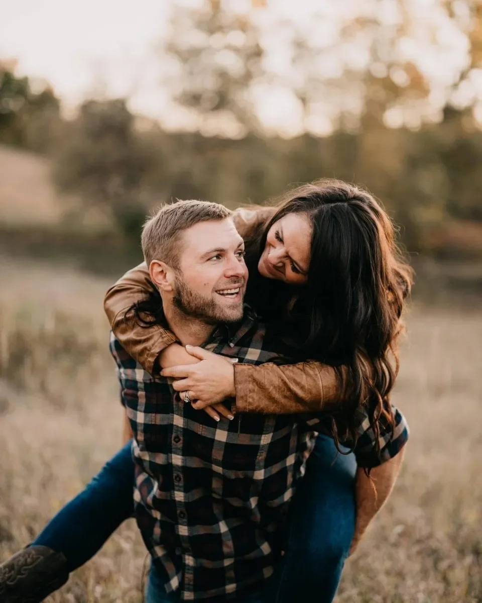 A couple outdoors, the woman is on the man's back, both smiling and laughing, during sunset with blurred trees in the background.