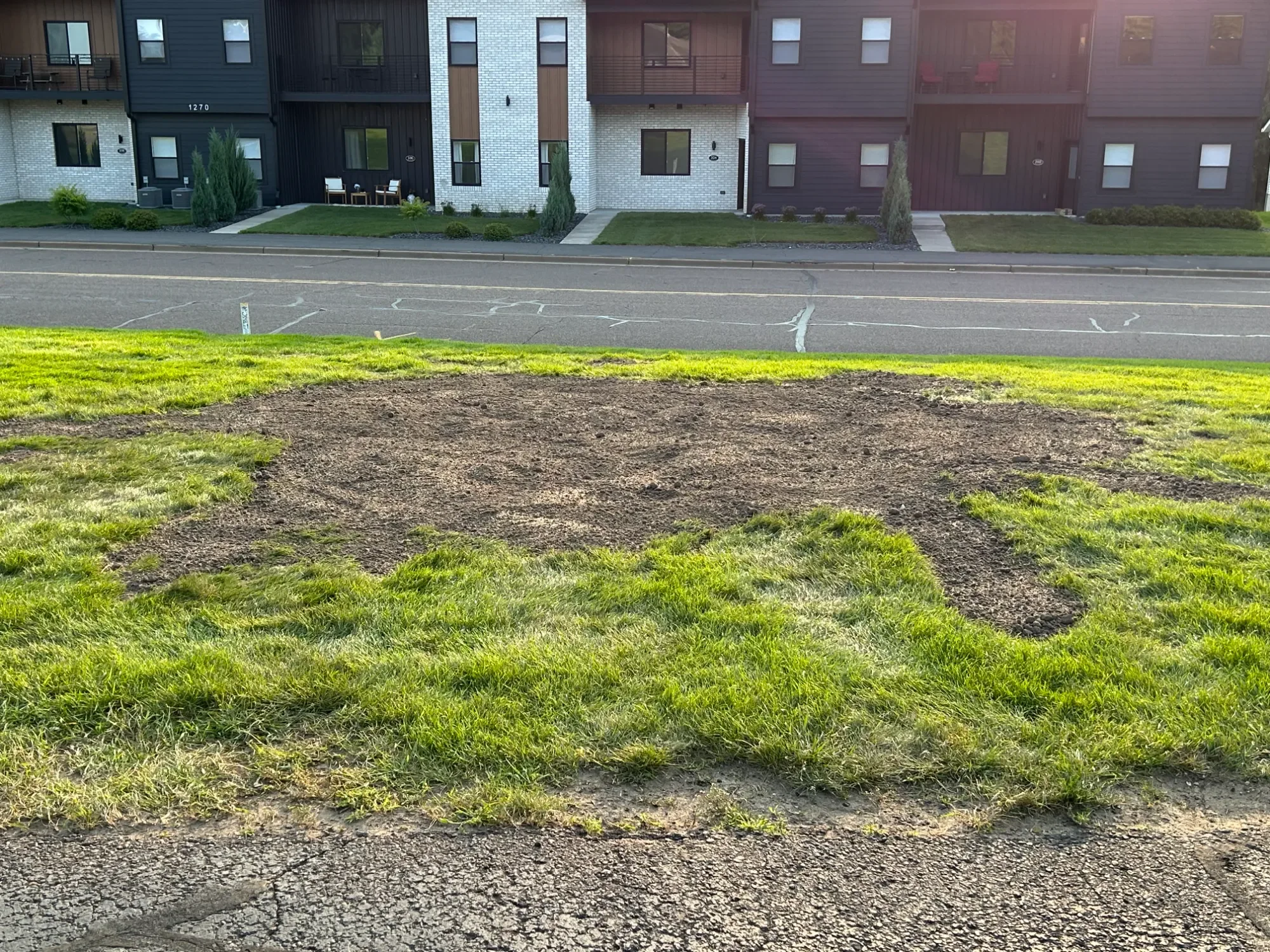 A patch of dirt and grass in front of a parking lot and modern apartment building with multiple windows and balconies.