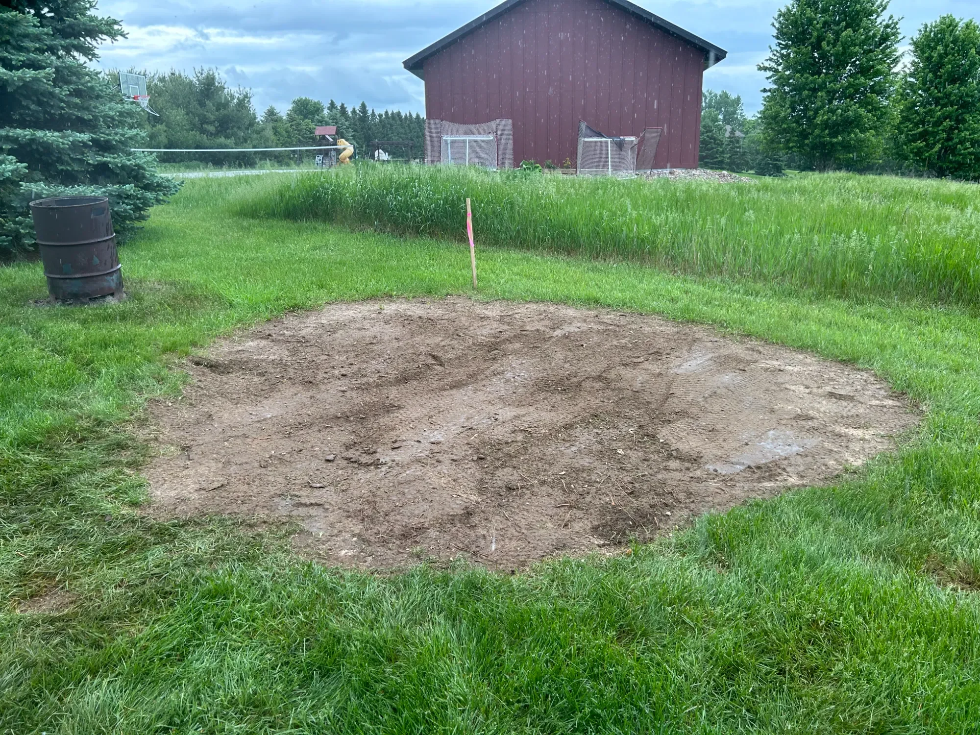 A patch of disturbed soil in a grassy backyard with a red shed in the background, surrounded by green trees and grass, cloudy sky, and some sports equipment.