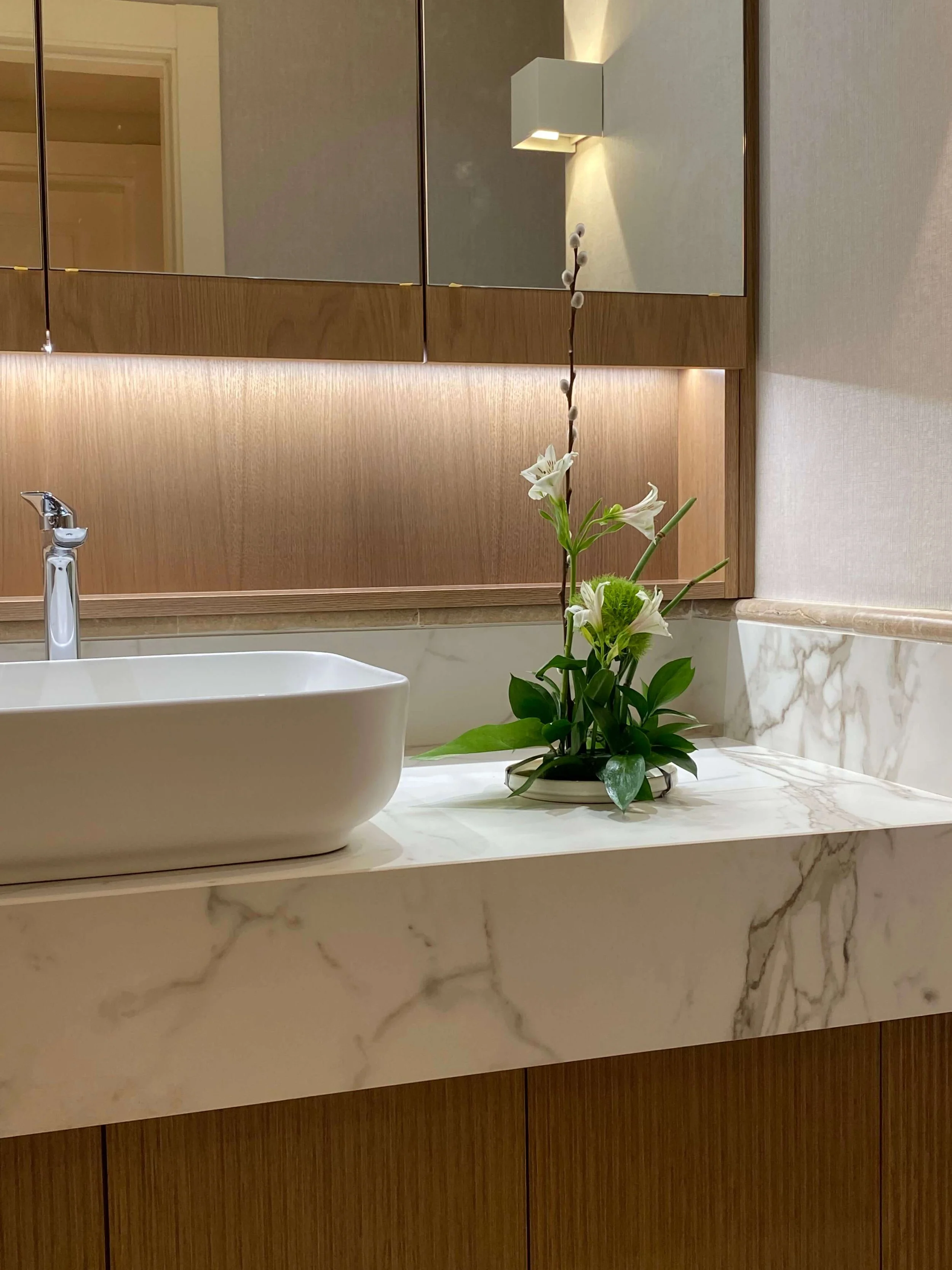 A bathroom vanity with a white vessel sink, a chrome faucet, and a flower arrangement with white flowers and greenery on a marble countertop. In the background, a wooden cabinet and mirrored doors with a wall-mounted light fixture are visible.