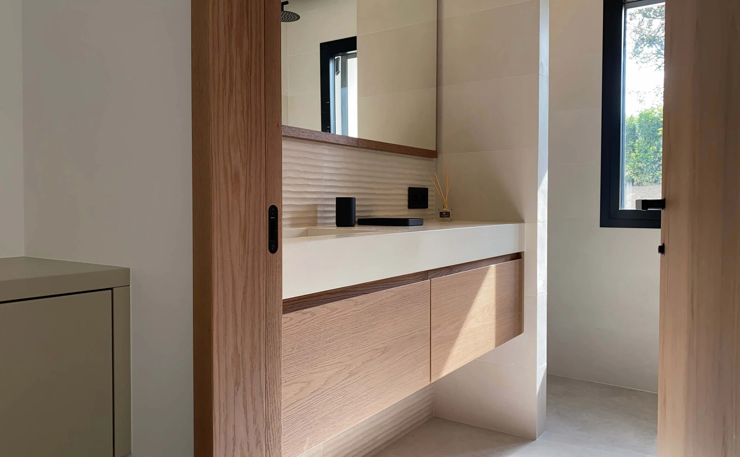 Minimalist bathroom vanity with a large mirror, a floating wooden cabinet, a textured beige backsplash, and a small reed diffuser. Natural light from a window illuminates the space.