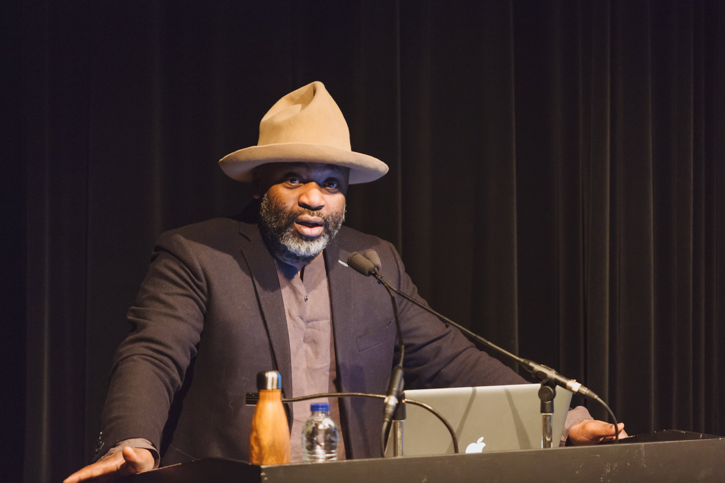 A man with a grey beard wearing a grey hat and dark blazer giving a speech behind a podium with a microphone, a water bottle, and a laptop, on a stage with black curtains.