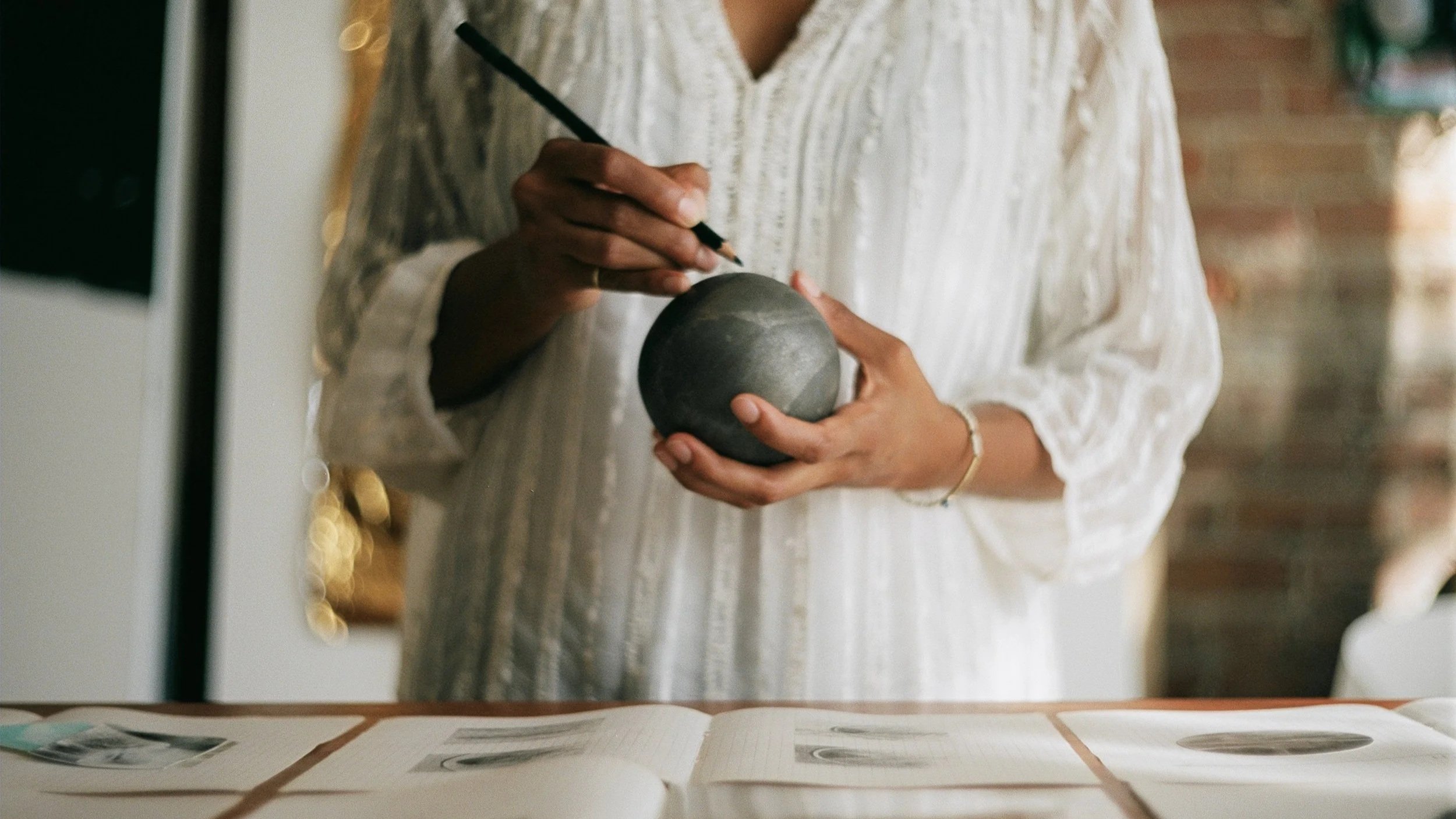 A person in a white, textured blouse is holding a black spherical object and writing or drawing on it with a black pen, in a room with warm lighting and brick wall in the background, with photographs laid out on a table in front.