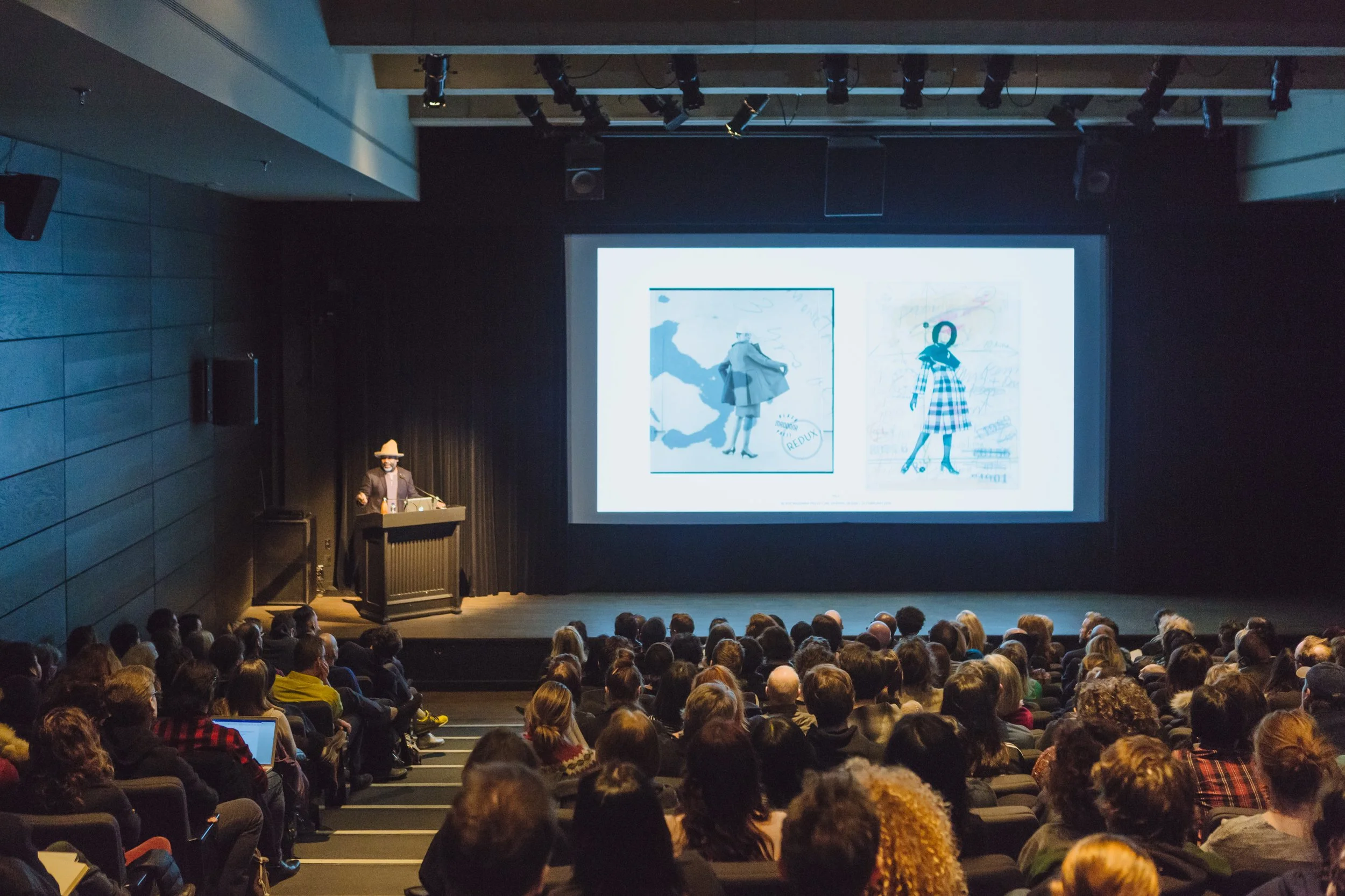 A person giving a presentation on stage in a darkened auditorium with a large screen behind them displaying two images of fashion sketches.