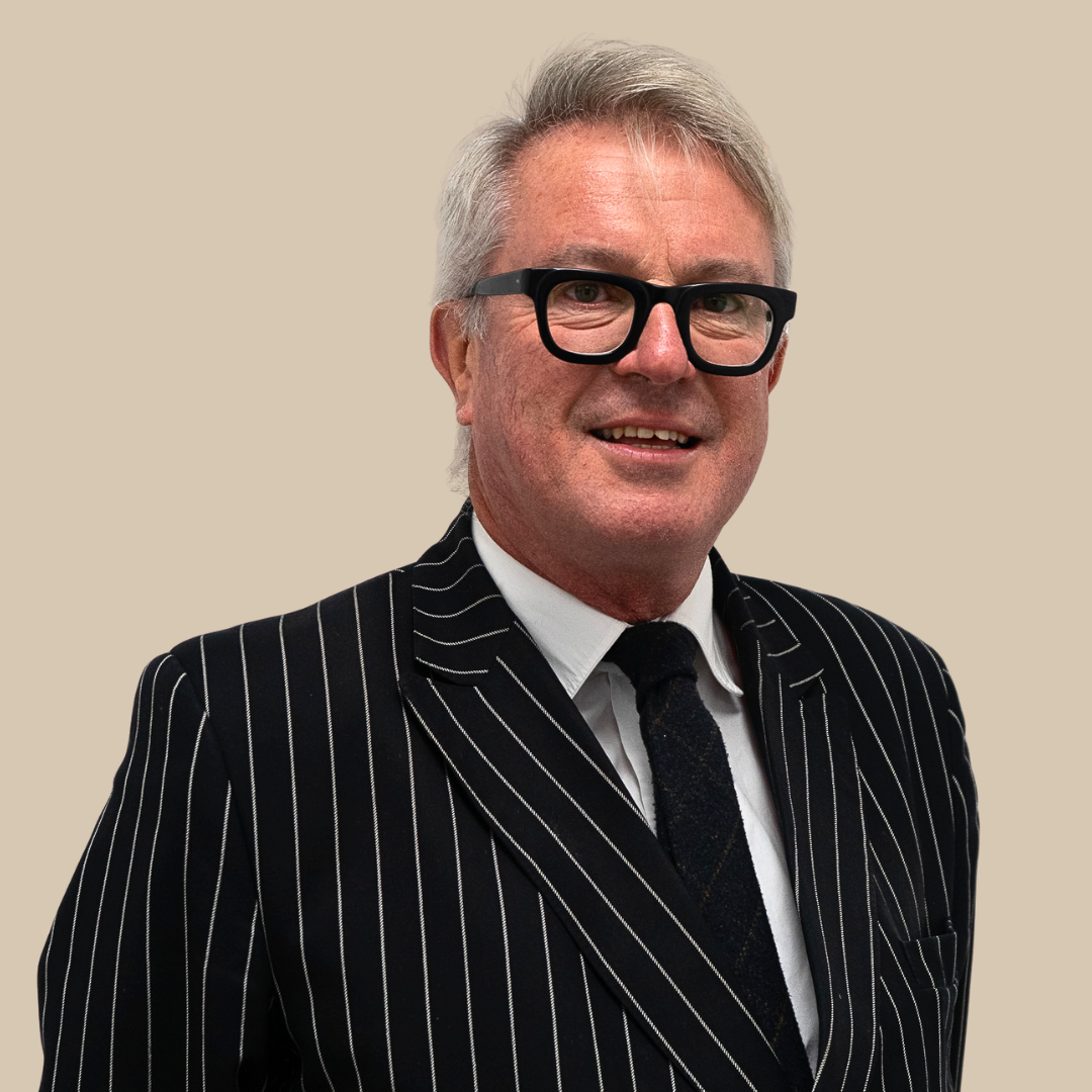 A middle-aged man with gray hair and black glasses, wearing a black and white pinstripe suit, white shirt, black tie, smiling against a plain beige background.