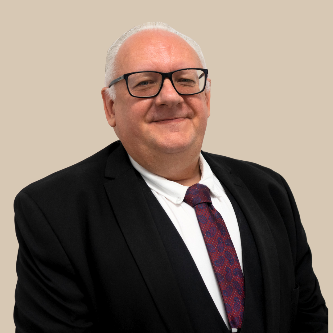 Portrait of a smiling middle-aged man with white hair and glasses, wearing a black suit, white shirt, and a red patterned tie, against a plain beige background.