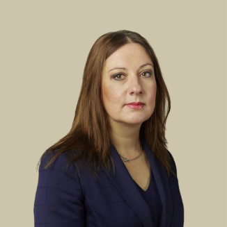 A woman with shoulder-length brown hair wearing a navy blazer and a necklace, posing against a beige background.