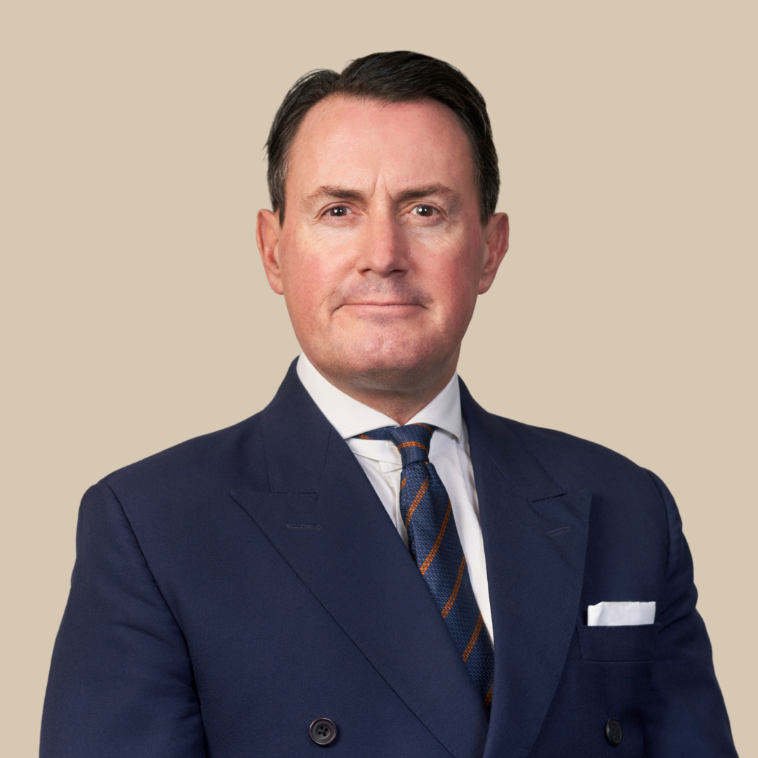 A middle-aged man dressed in a dark navy suit, white shirt, and a diagonally striped tie, posing against a beige background.