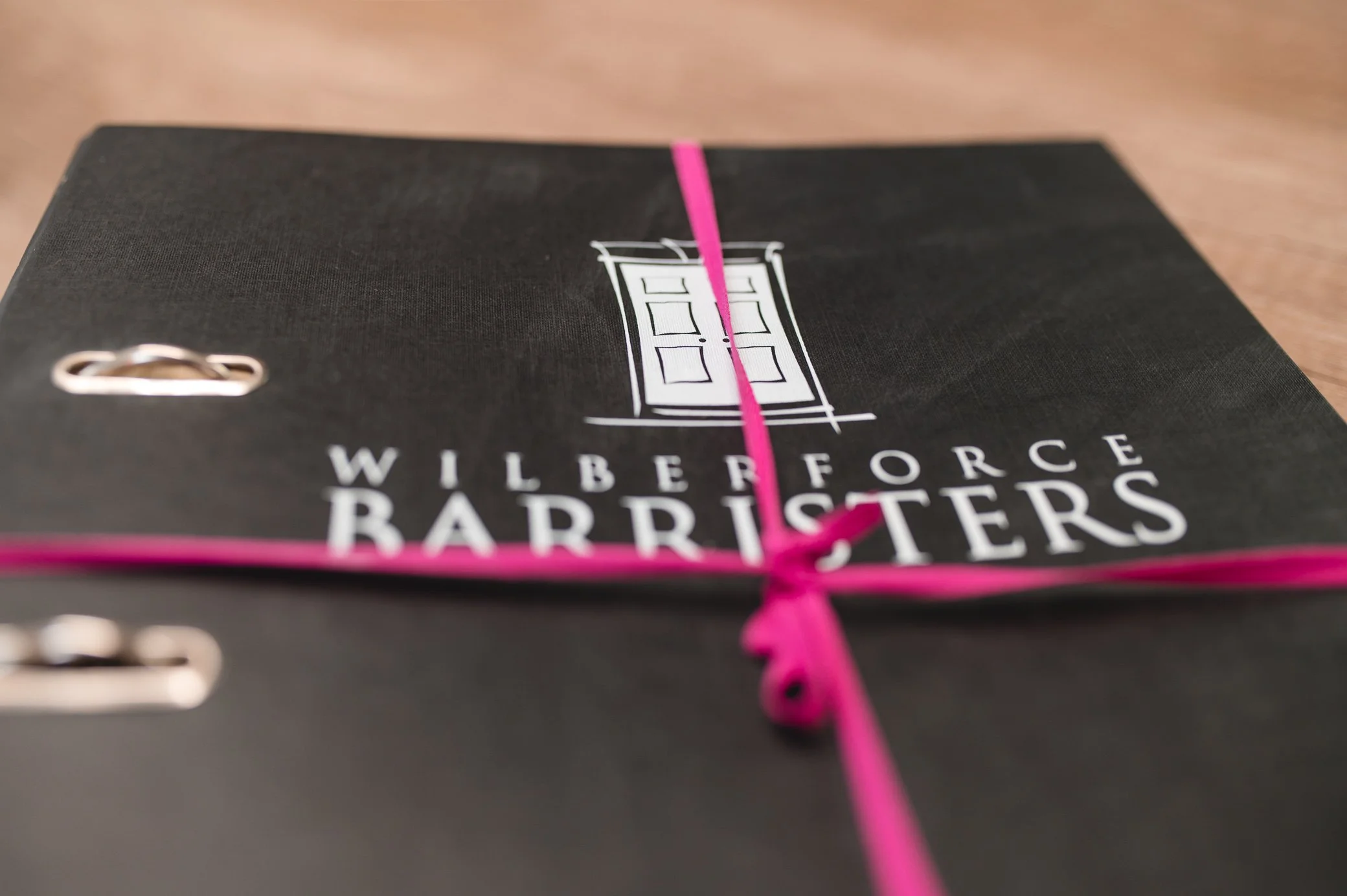 A black folder or binder with the Wilberforce Barristers logo and name on it, tied with a pink ribbon, lying on a wooden surface.