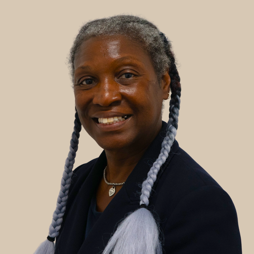 A smiling woman with dark skin, gray and black braided hair, wearing a dark blazer and a beaded necklace with a leaf charm.