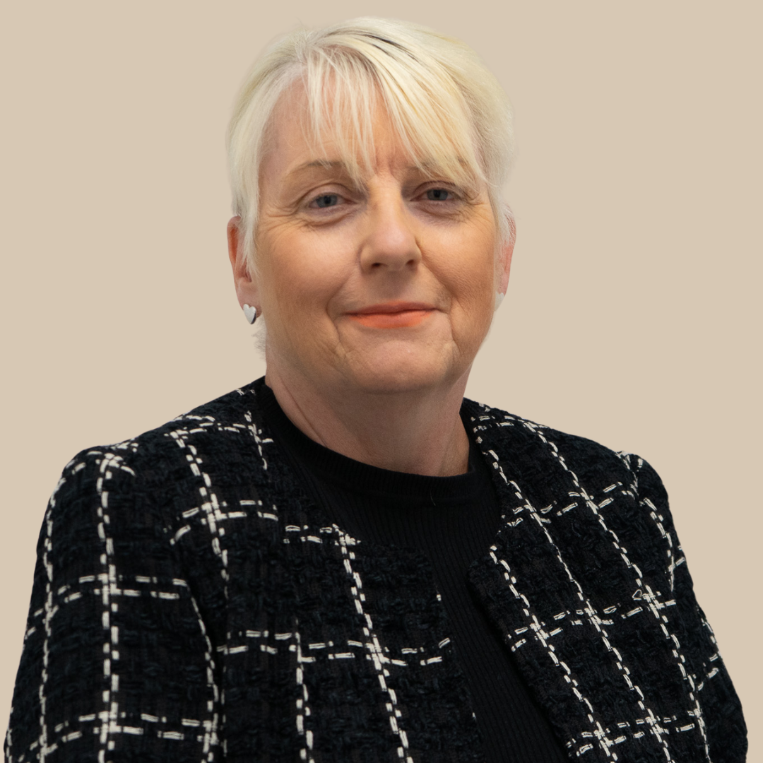 A portrait of an older woman with short blonde hair, wearing a black and white checked jacket and earrings, against a plain background.