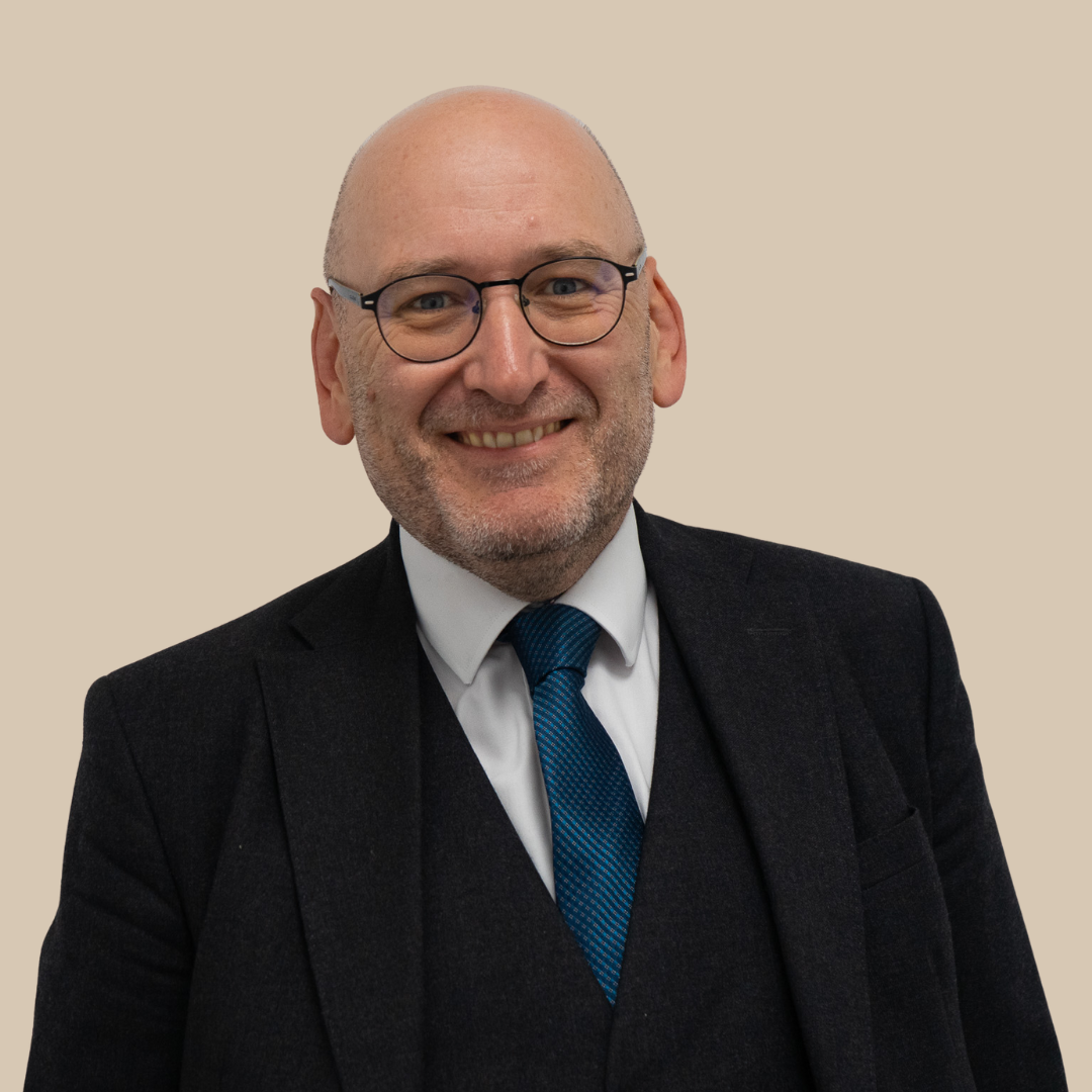 A middle-aged man with glasses smiling, wearing a black suit, white shirt, and blue tie, standing against a plain beige background.