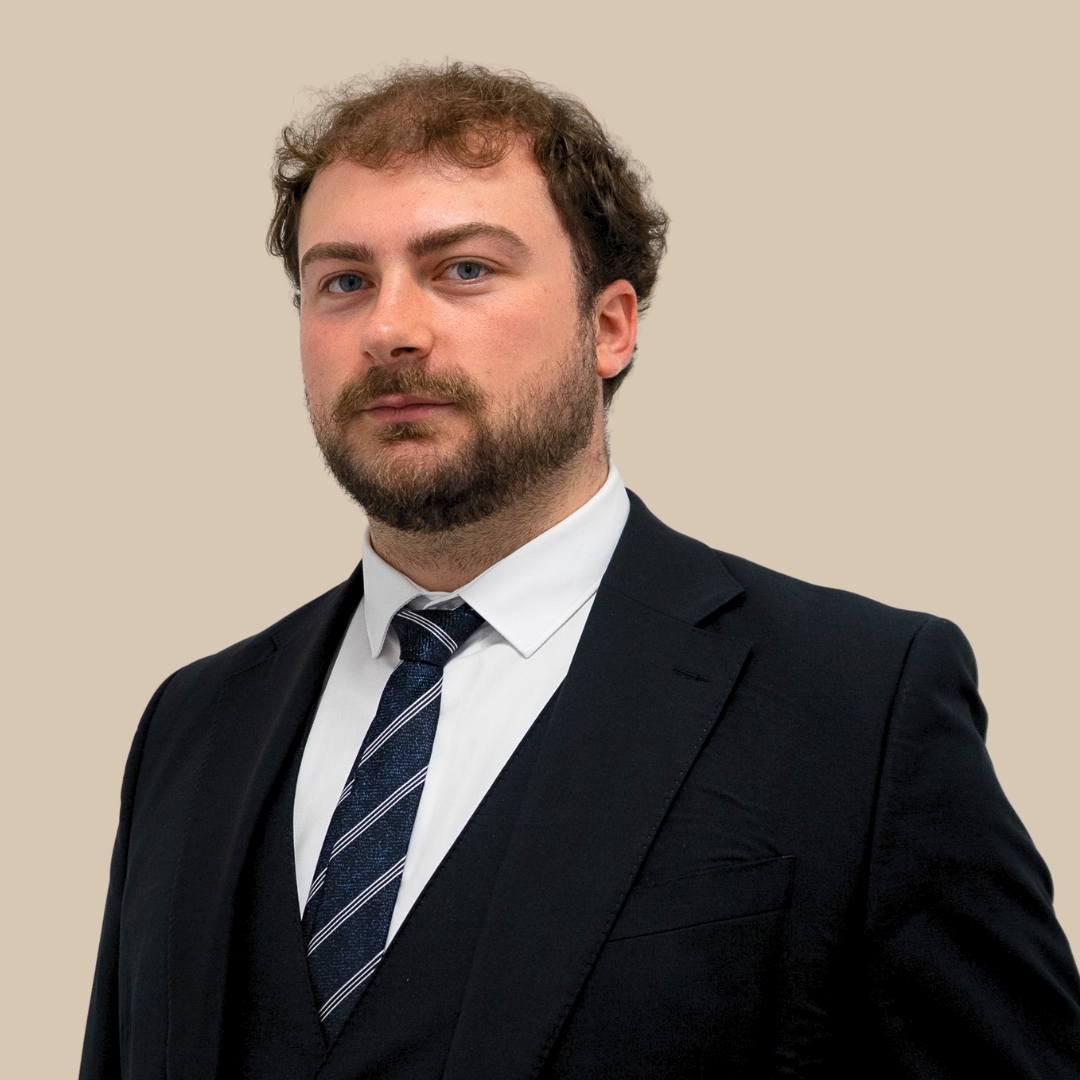 A man in a dark suit and tie with a white shirt, looking confidently at the camera against a plain beige background.