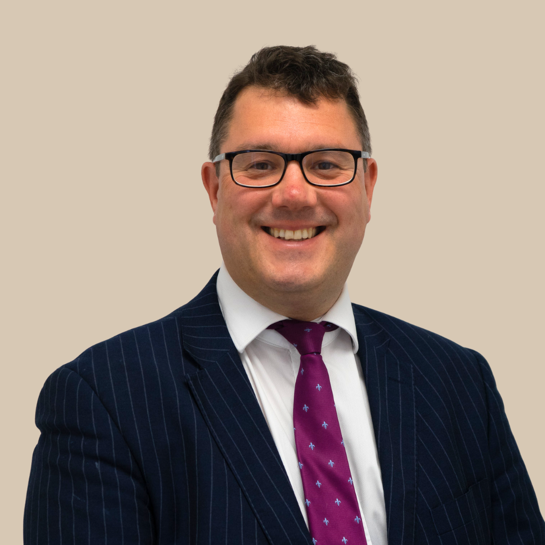 A smiling man wearing glasses, a navy pinstripe suit, a white shirt, and a purple tie with small floral patterns, against a beige background.