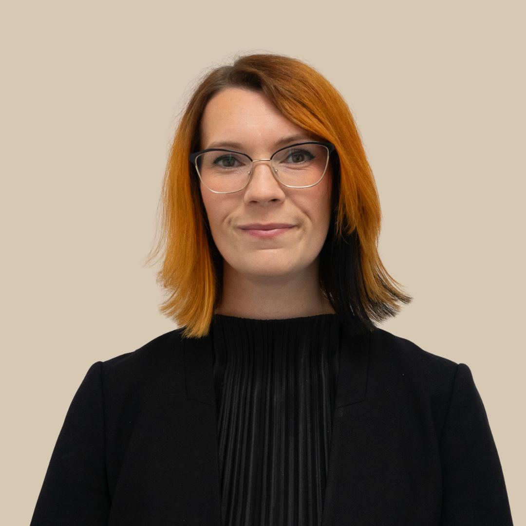 A woman with shoulder-length orange and black hair, wearing glasses, and dressed in a black top, standing against a plain beige background.