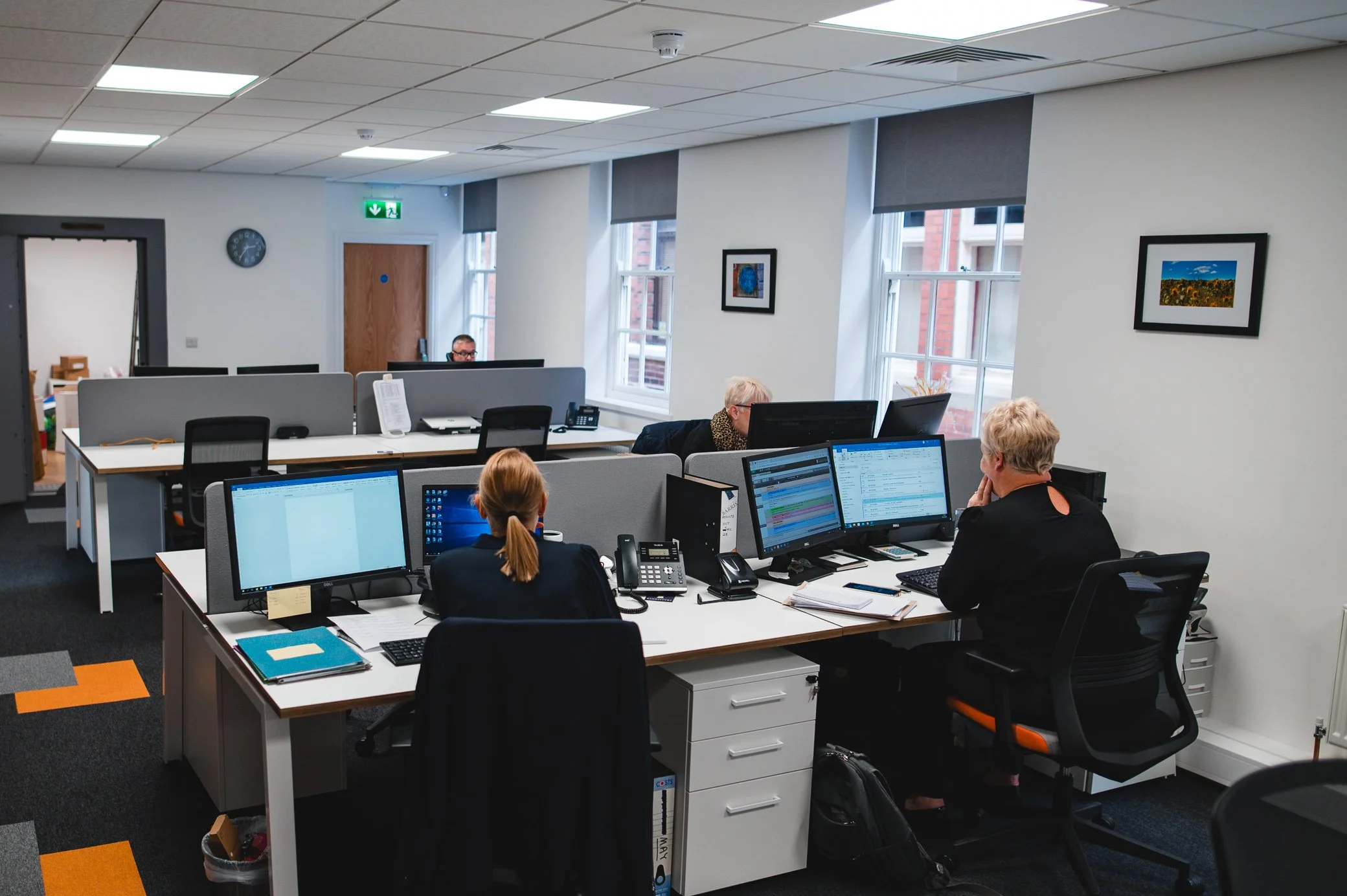 Office with multiple employees working at desks with computers, offices, and artwork on white walls.