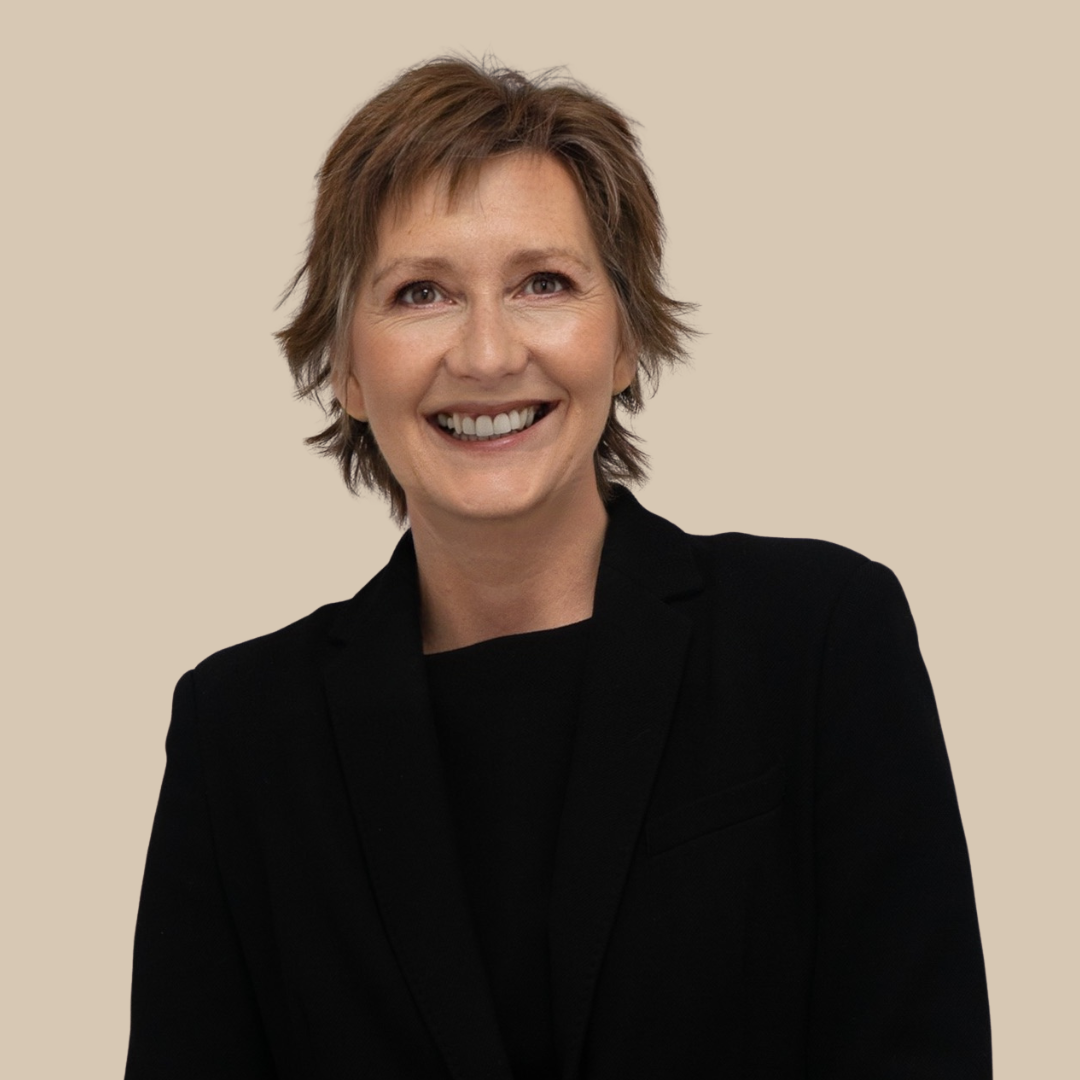 A woman with short, light brown hair smiling and wearing a black blazer against a beige background.