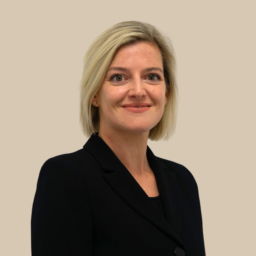 A woman with shoulder-length blonde hair wearing a black blazer, smiling, against a plain beige background.