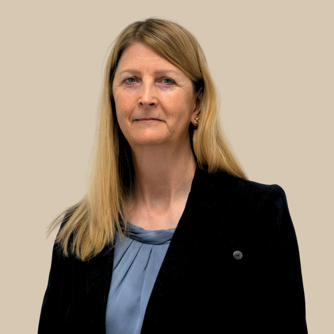 A middle-aged woman with long blonde hair, wearing a black blazer and a gray blouse, standing against a beige background.