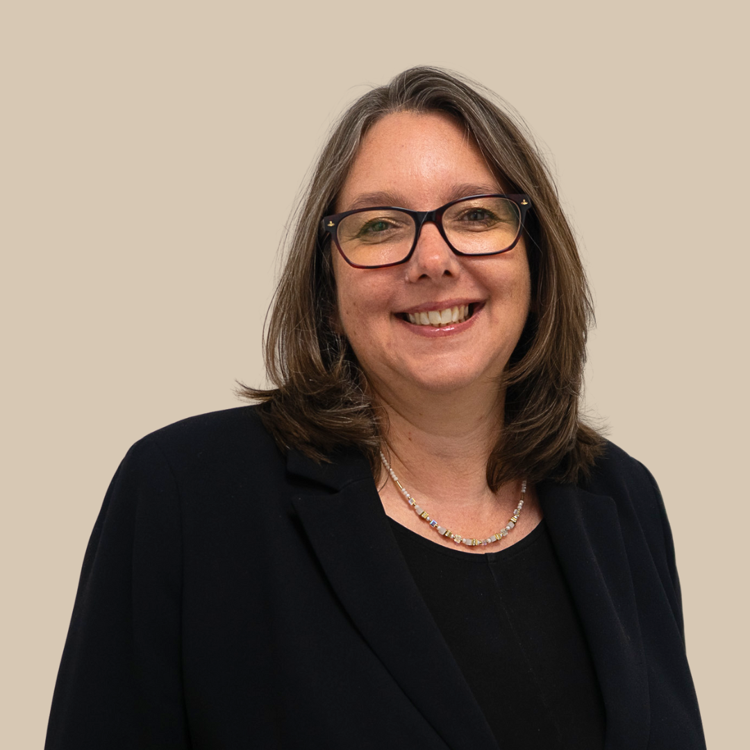 A woman wearing glasses, smiling, with shoulder-length brown hair, dressed in a black blazer and a necklace, against a beige background.
