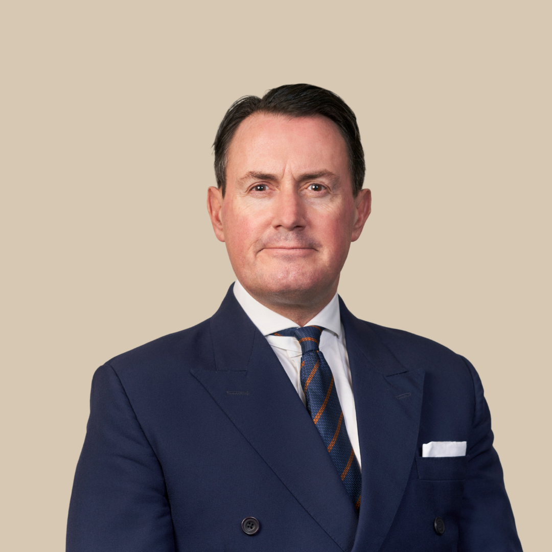 A man in a navy blue suit, white shirt, and a striped tie, standing against a beige background.