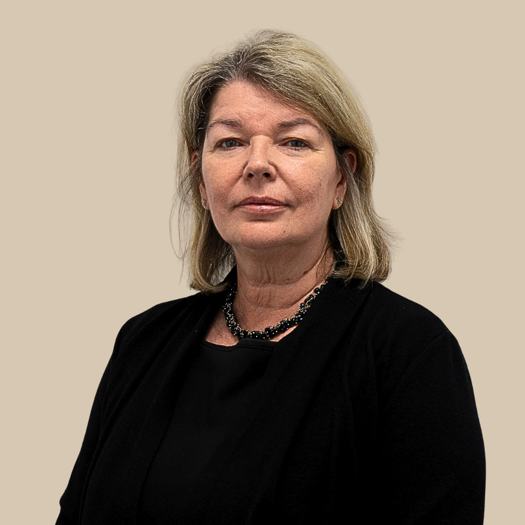 A middle-aged woman with shoulder-length blonde hair, wearing a black top and necklace, looking directly at the camera with a neutral expression, standing against a plain beige background.