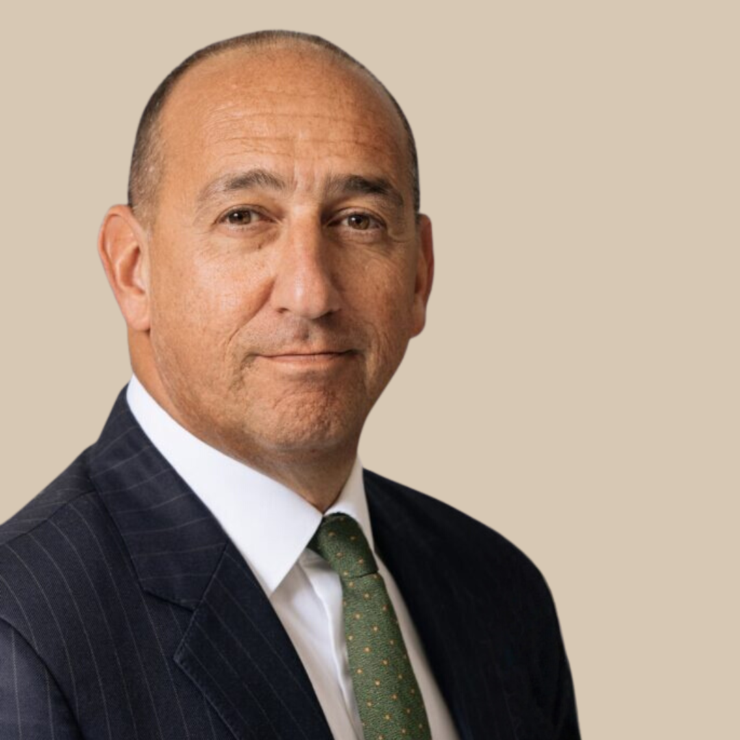 A middle-aged man with a serious expression, dressed in a dark pinstripe suit, white shirt, and green tie, against a plain beige background.
