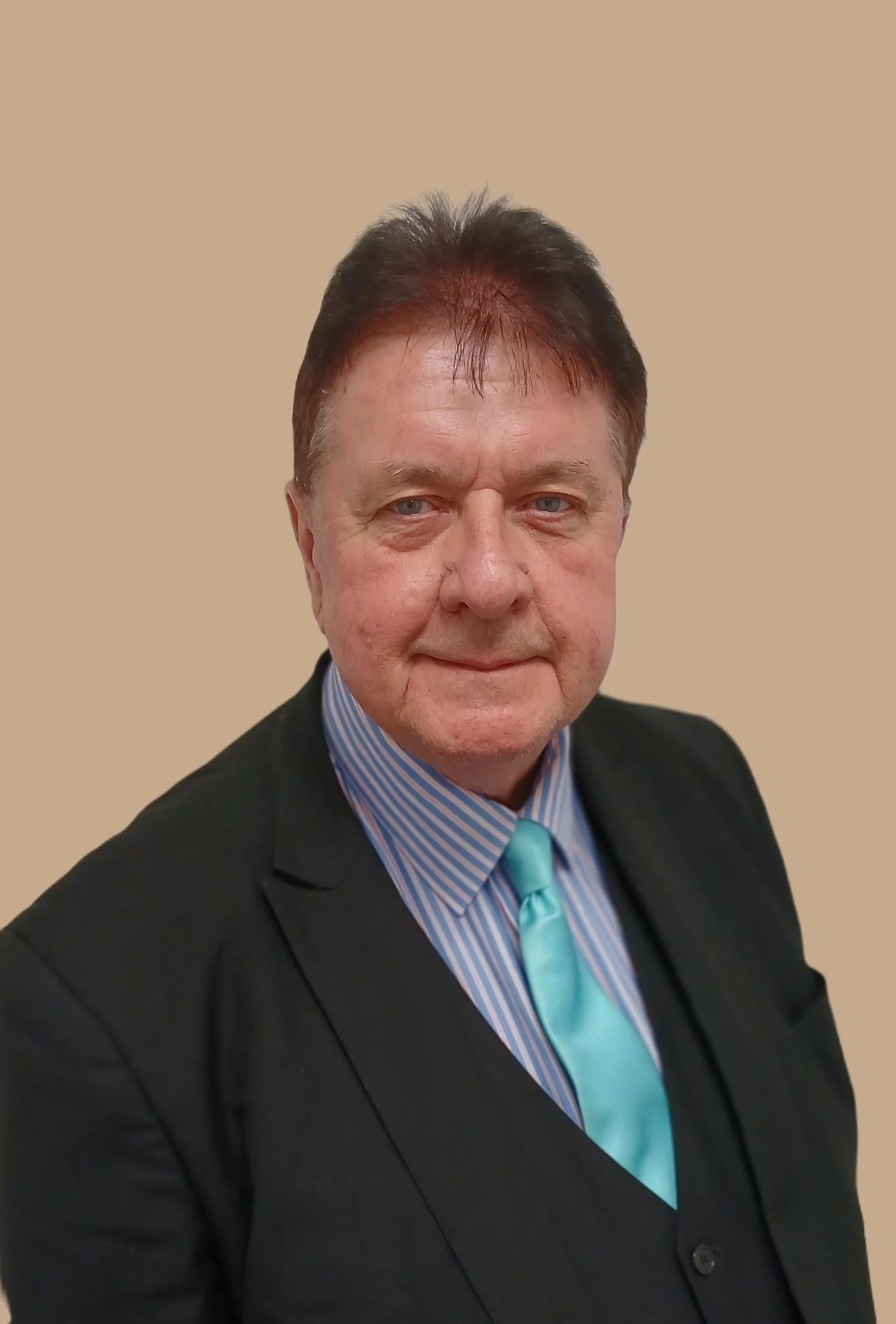 A man in a black suit with a white shirt and yellow striped tie, standing against a plain beige background.