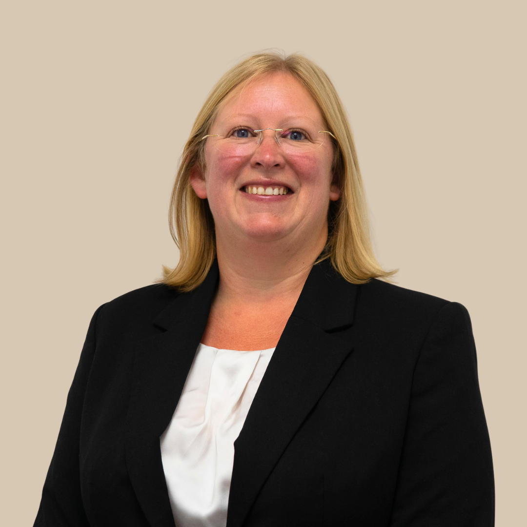 A woman with blonde hair and glasses, dressed in a black blazer and white blouse, smiling against a beige background.