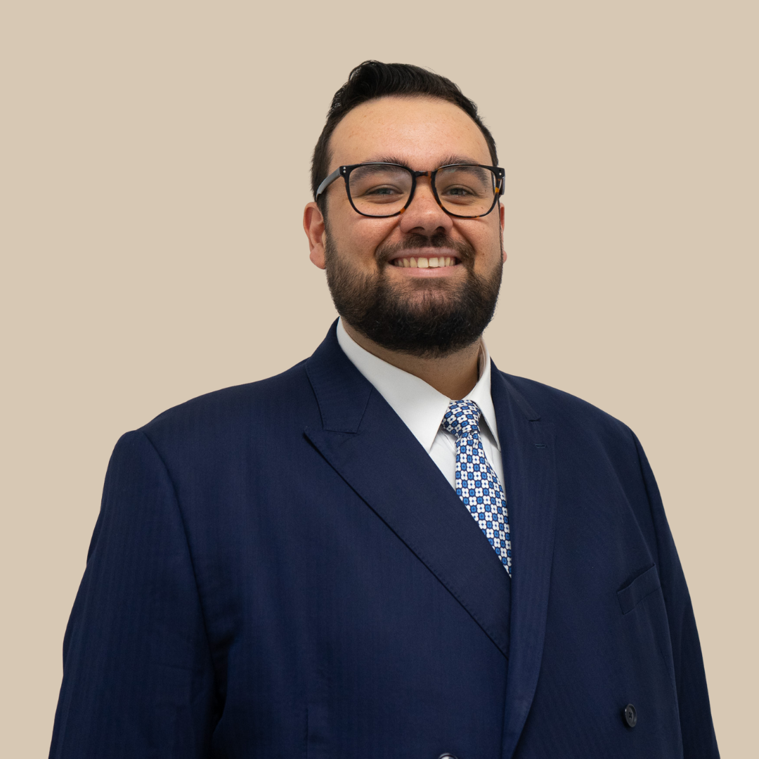 A man wearing glasses, a navy blue suit, white shirt, and patterned tie, smiling against a beige background.