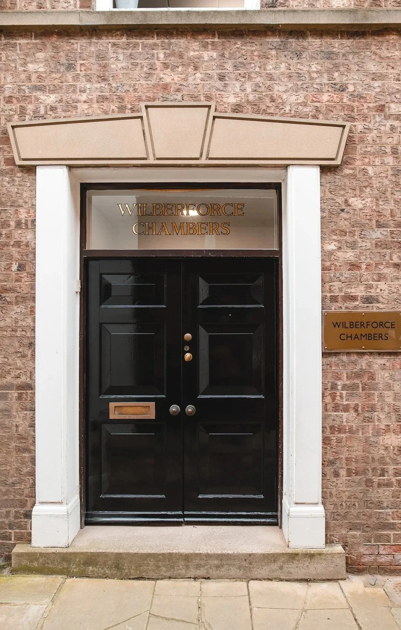 Black double door entrance with glass top section, framed by white trim and a brick building. Brass plaque and gold lettering on the glass read 'Wilberforce Chambers'.