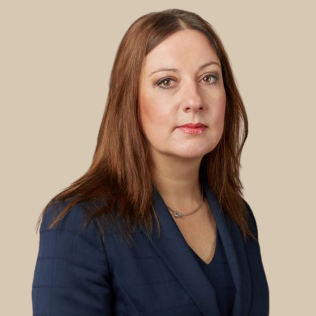 A woman with medium-length reddish-brown hair wearing a navy blazer and a subtle necklace, standing against a plain beige background.