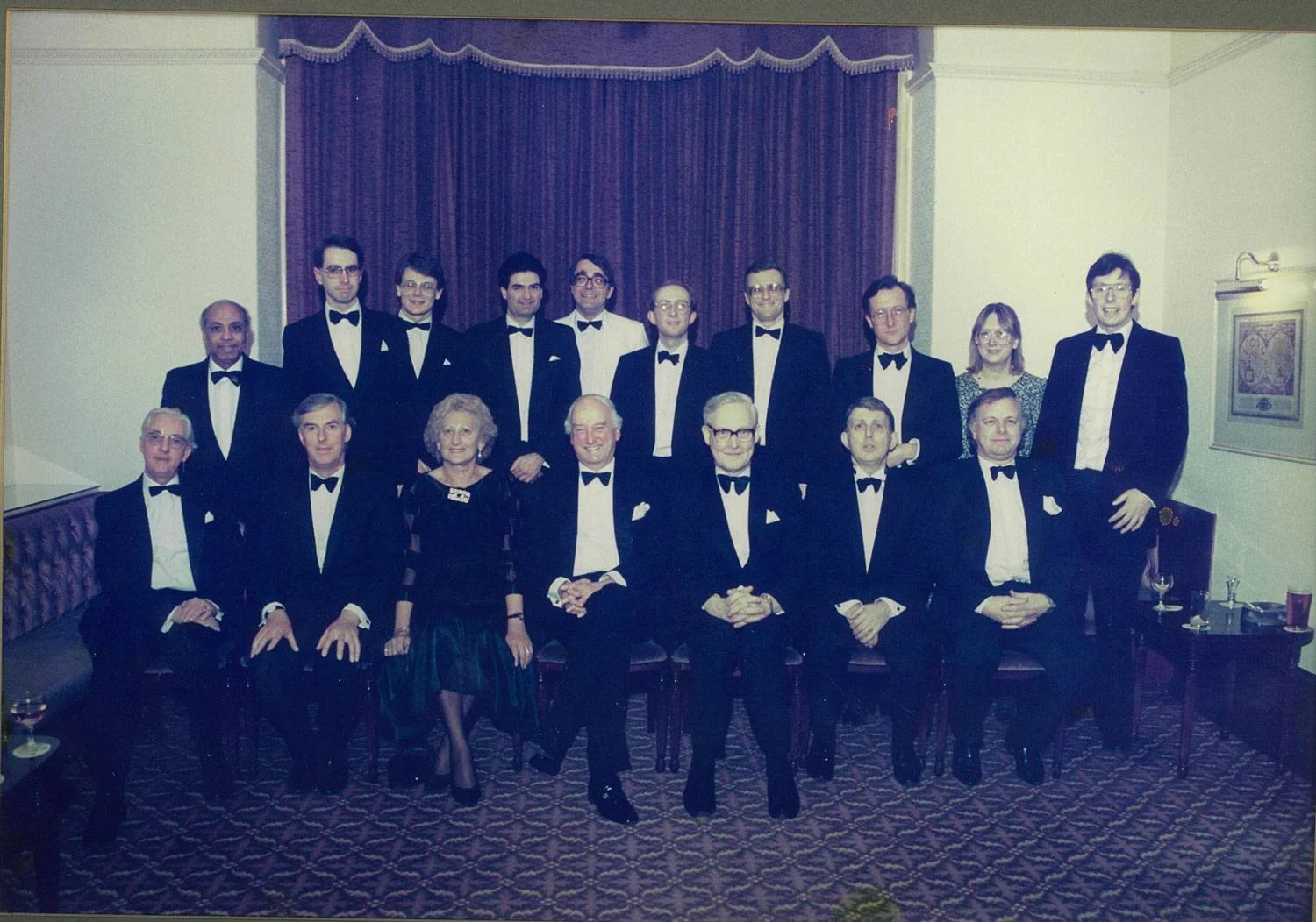 A group of 16 people dressed in formal tuxedos and evening dresses pose for a photograph in a decorated room with a purple curtain backdrop.