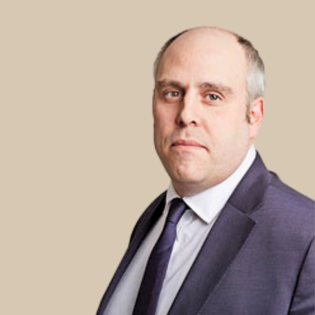 A man wearing a dark suit, white shirt, and purple tie against a beige background.