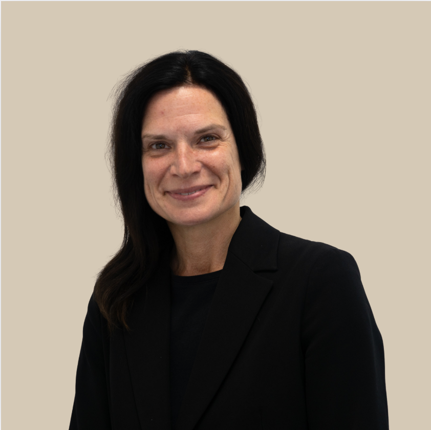 A woman with black hair wearing a black blazer, smiling against a beige background.