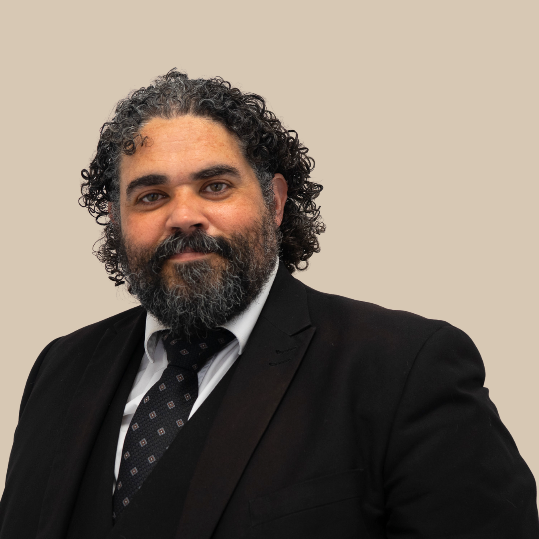 A man with curly dark hair and a beard wearing a black suit, white shirt, and patterned black tie, standing against a plain beige background.