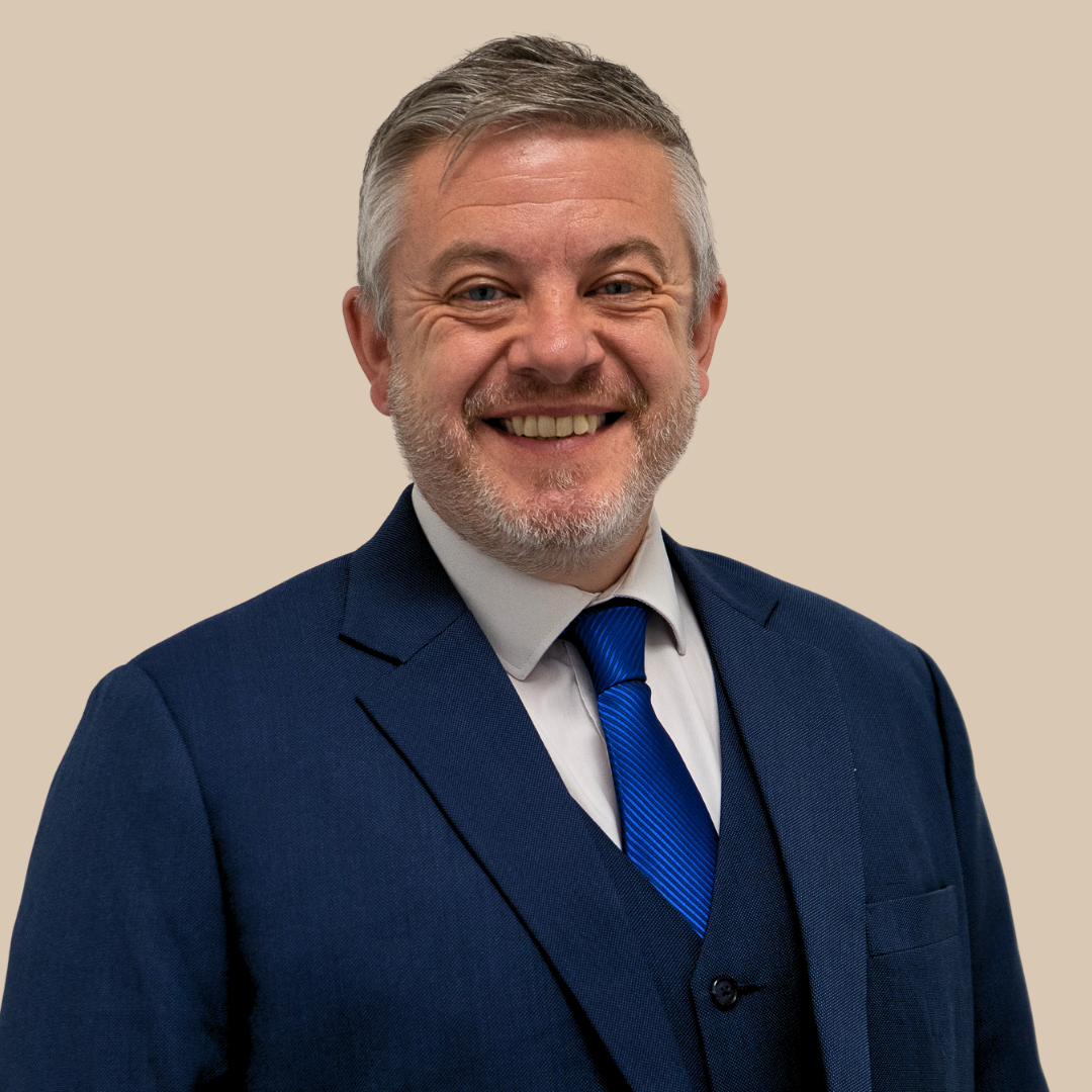 A smiling middle-aged man with gray hair and a beard, wearing a navy blue suit, white shirt, and blue striped tie, standing against a beige background.