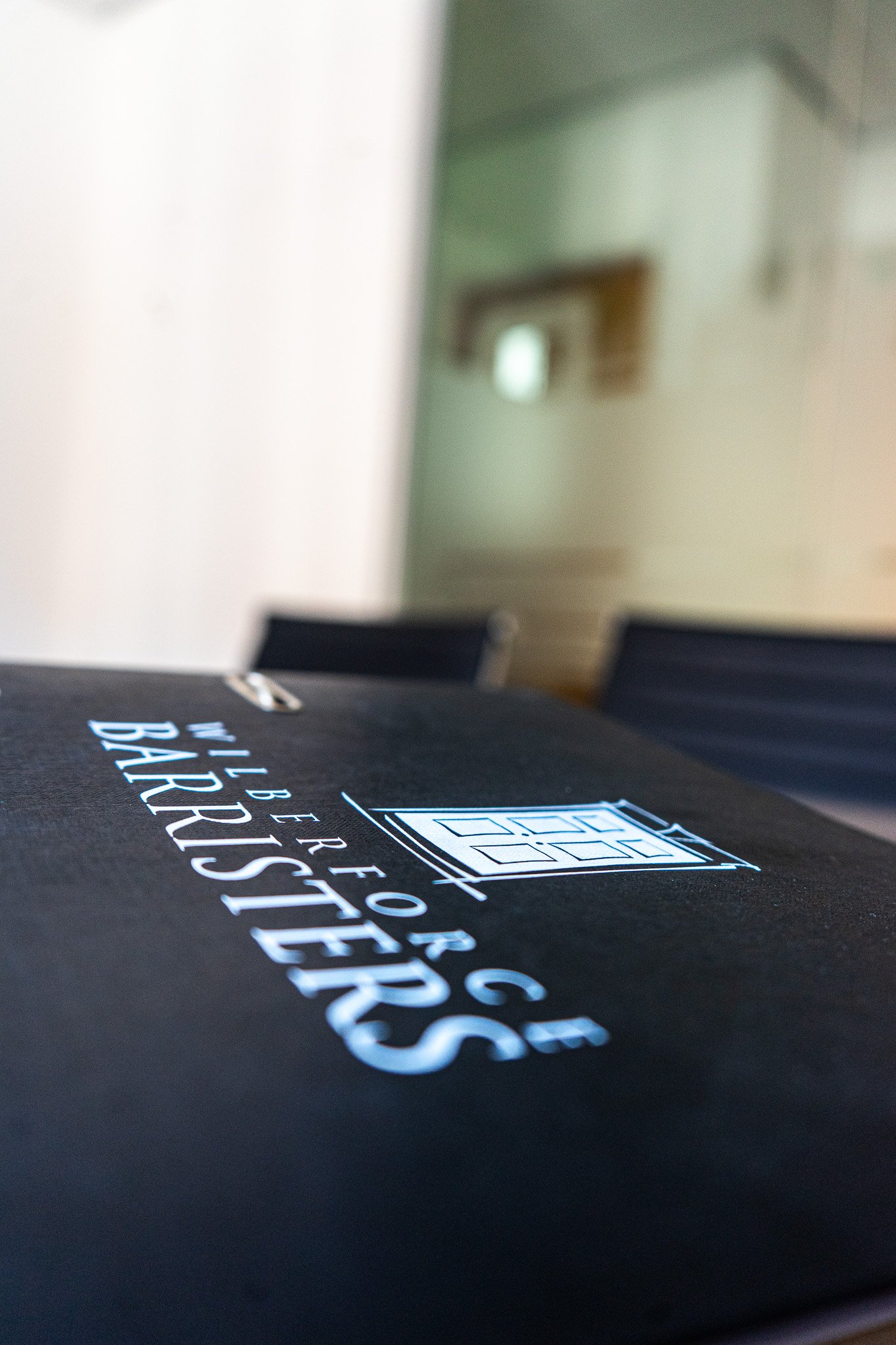 Close-up of a black folder or binder with the words 'Walter For G' and 'BARRISTERS' printed on it, placed on a table in a room with chairs and a beige wall in the background.