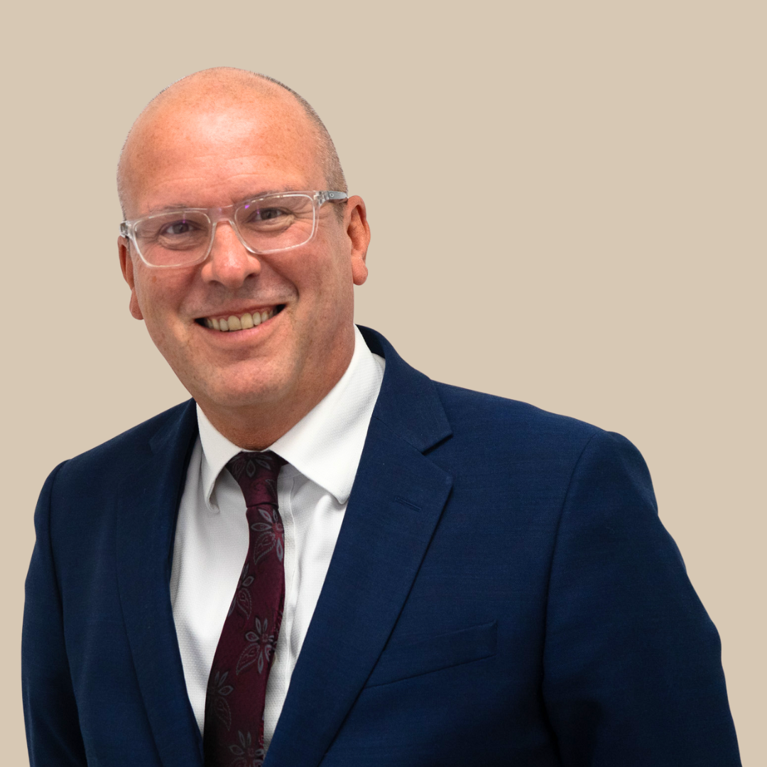 A man in a dark blue suit, white shirt, and maroon tie smiling at the camera, against a beige background.