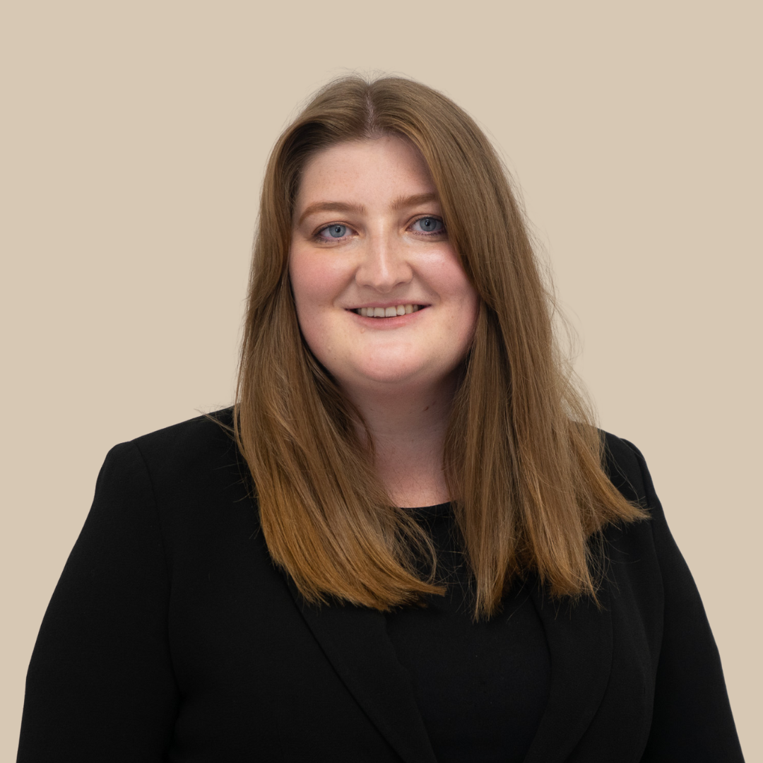 A woman with shoulder-length light brown hair and blue eyes, wearing a black blazer, smiling against a beige background.