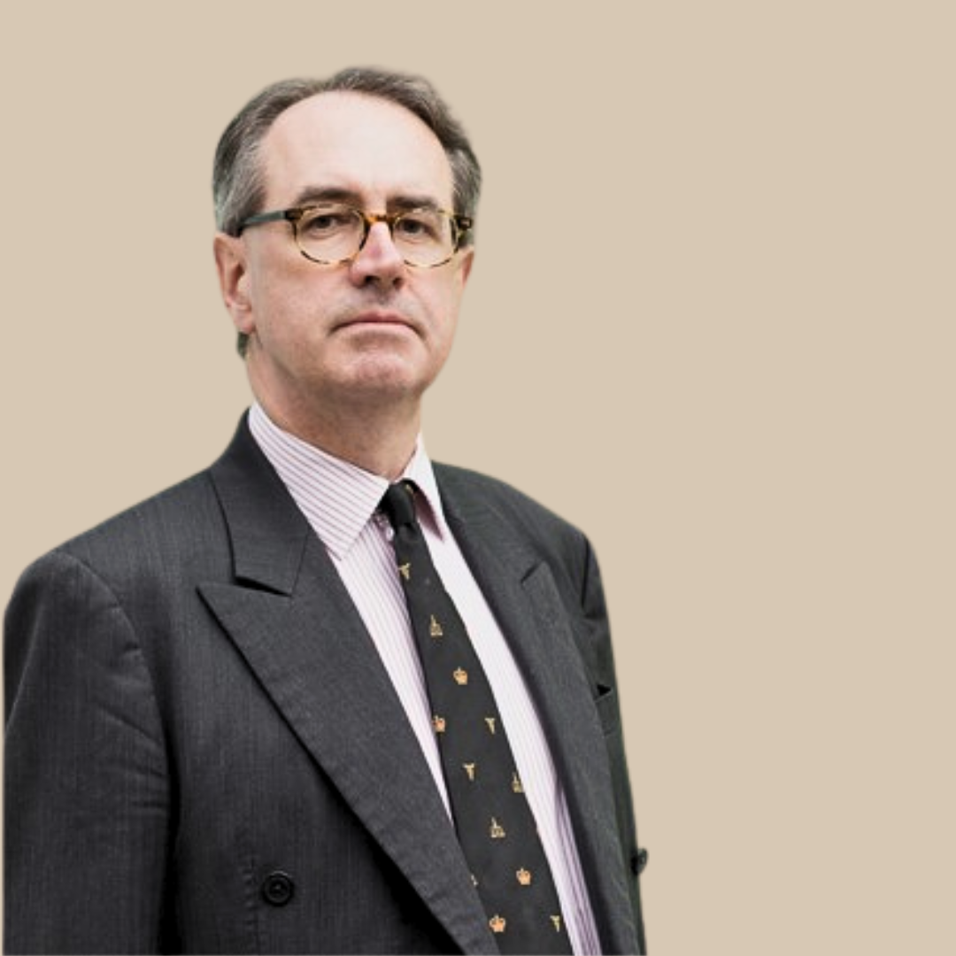 A middle-aged man wearing glasses, a dark gray suit, a pink striped shirt, and a black tie with small gold icons, standing against a beige background.
