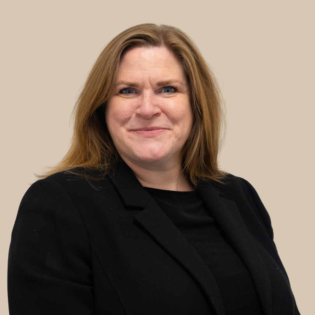 A woman with shoulder-length reddish-brown hair wearing a black blazer and black top, smiling slightly, against a beige background.