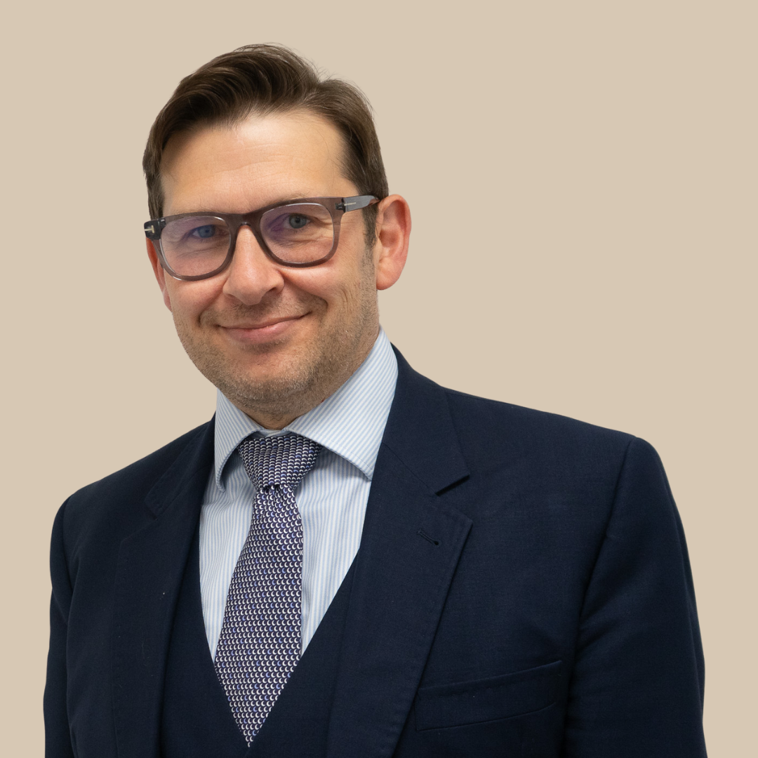 A man wearing a navy blue suit, light blue dress shirt, patterned tie, and glasses. He has short brown hair and is smiling against a beige background.