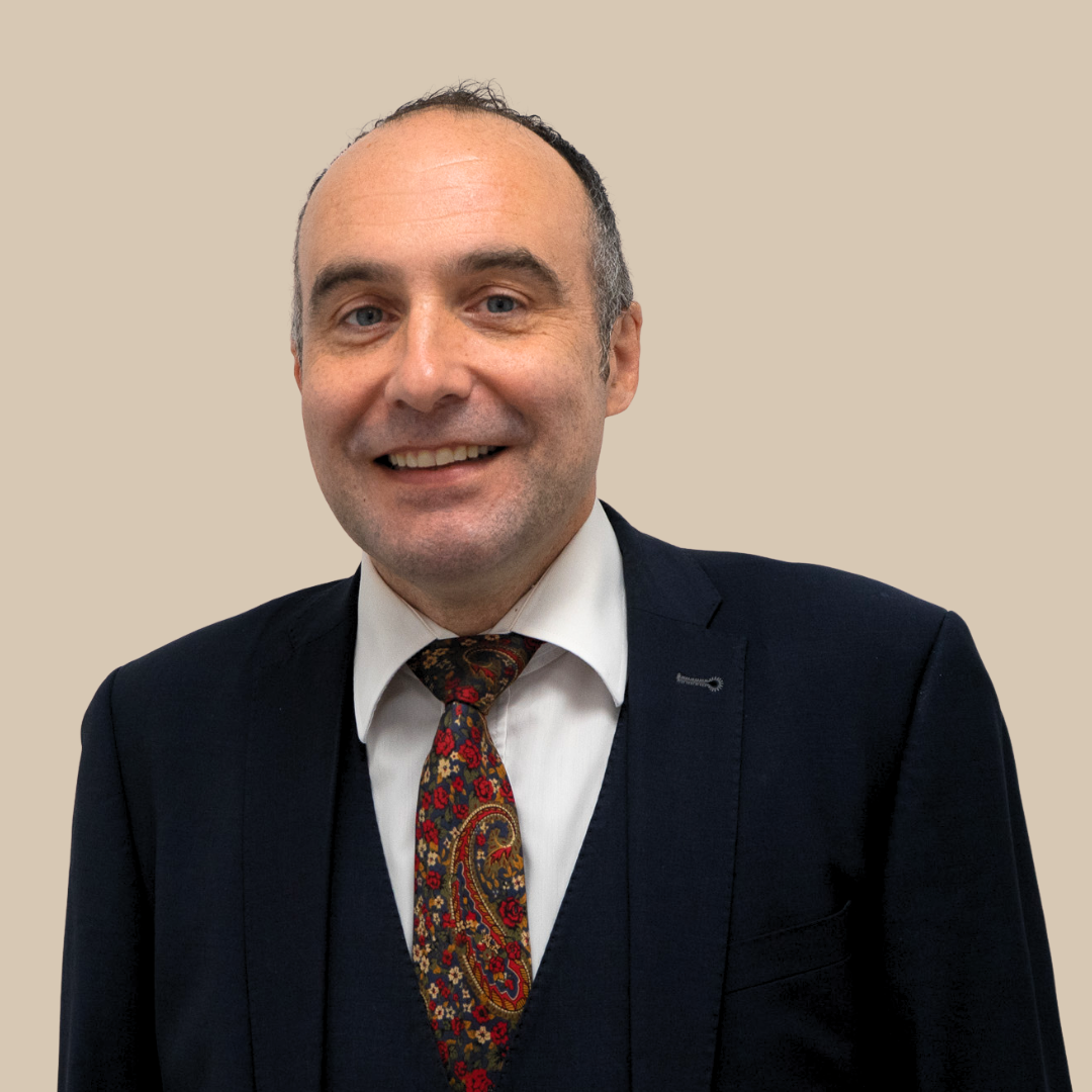 A smiling man in a dark suit with a colorful paisley tie and a white shirt, standing against a plain beige background.