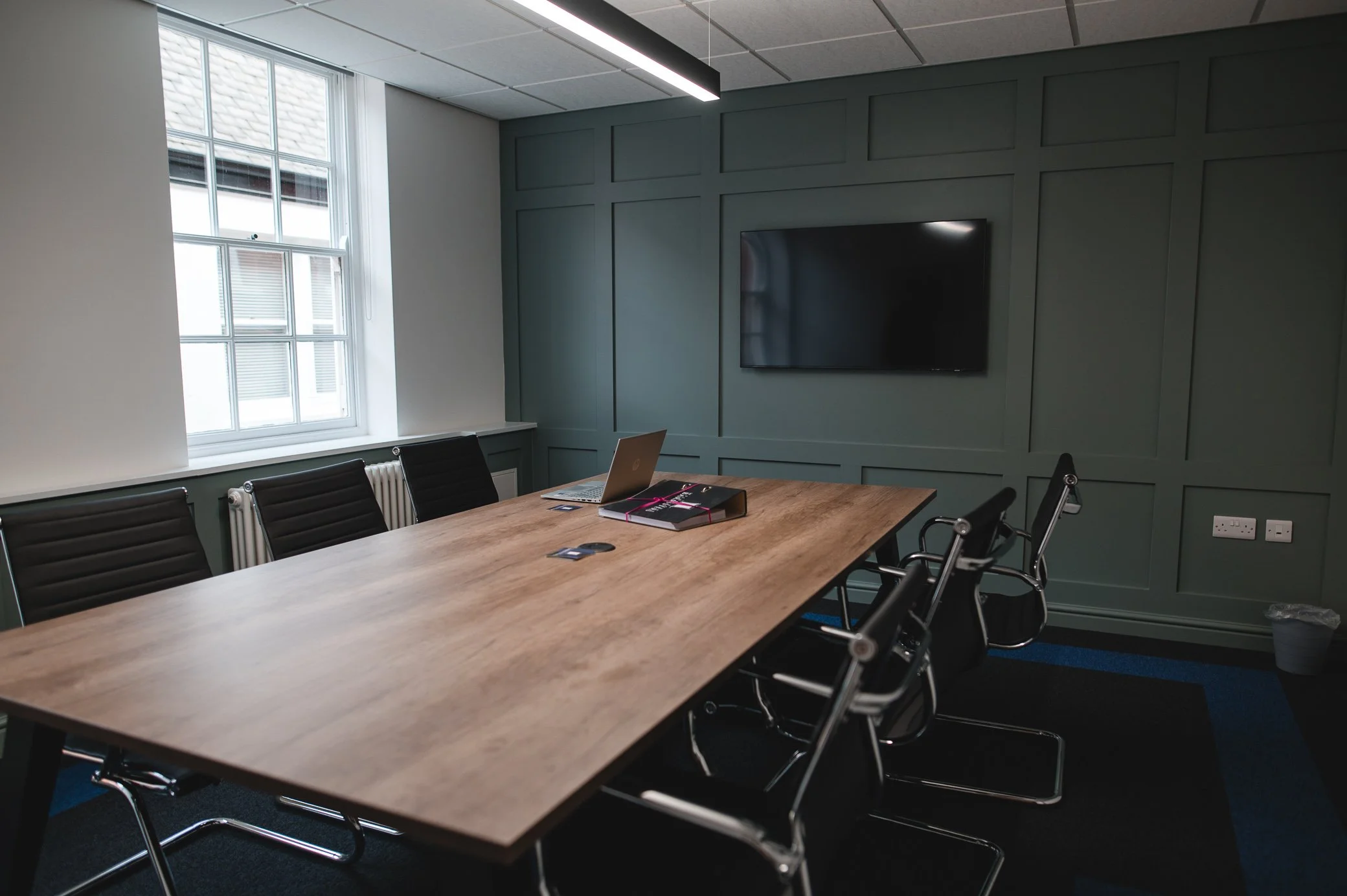 A modern conference room with a large wooden table, black leather chairs, a wall-mounted TV, and a laptop with open notebooks on the table. There is a window with white framing and a radiator beneath it, and the wall behind the TV features paneled wall design. Overhead lighting is mounted on the ceiling.